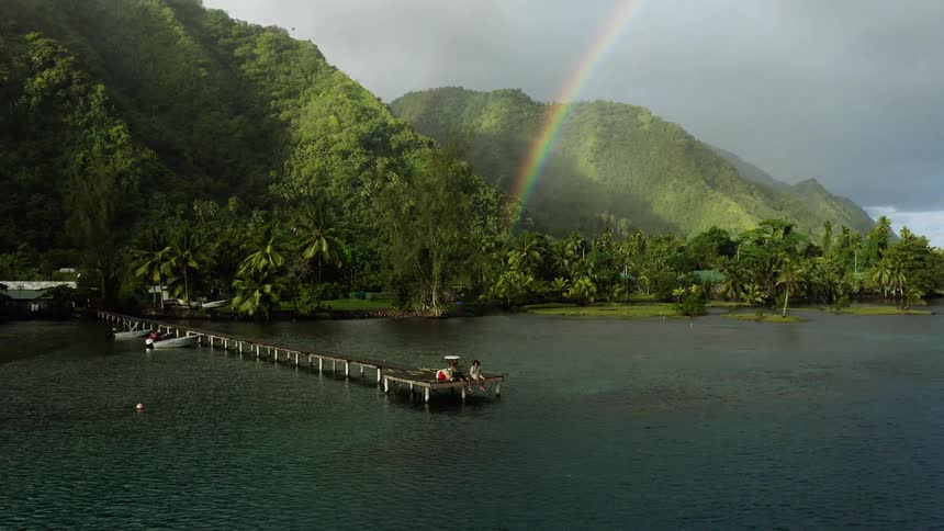 Rainbow arcs through the sky over a coastal forest while people sit at the end of the pier on an overcast misty day