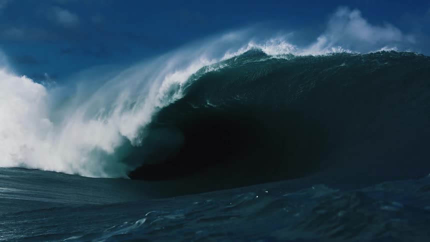 A massive ocean wave crests and breaks into a deep barrel creating a heavy wall of water and thick white spray against a dark blue sky and choppy sea surface