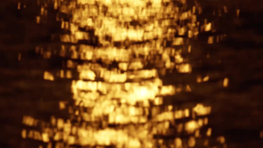 Golden sunlight reflects off the ocean surface in a shimmering trail of bokeh as the camera tilts upward from the sparkling waves to reveal a dark atmospheric sunset sky with thin clouds resting on the horizon