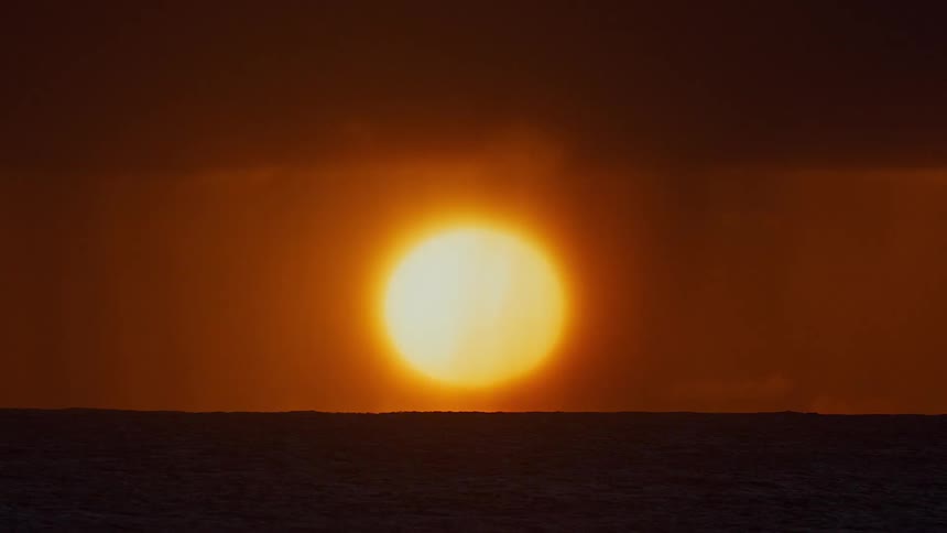 The glowing sun sets over a calm ocean on the horizon beneath a warm orange sky