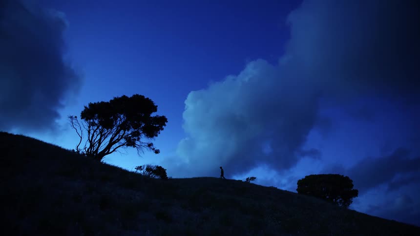 Silhouette of a man walking on a mountain hill as trees move in the wind with clouds overhead