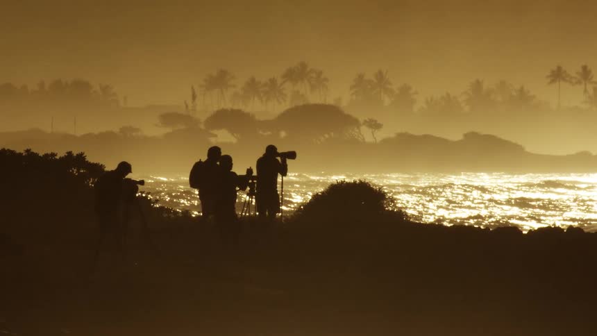Four photographers stand on a rocky shoreline silhouetted against a bright golden sunset while they point their cameras and long lenses toward the shimmering ocean waves and a distant palm tree lined coast