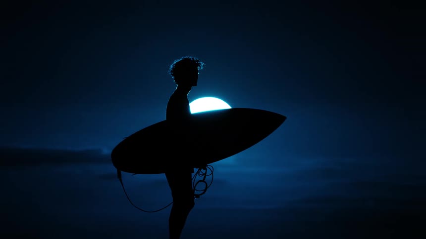 Silhouette of a surfer holding a board against a low moon over the sea at night