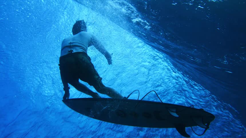 A surfer rides a wave underwater as sunlight filters through the blue ocean