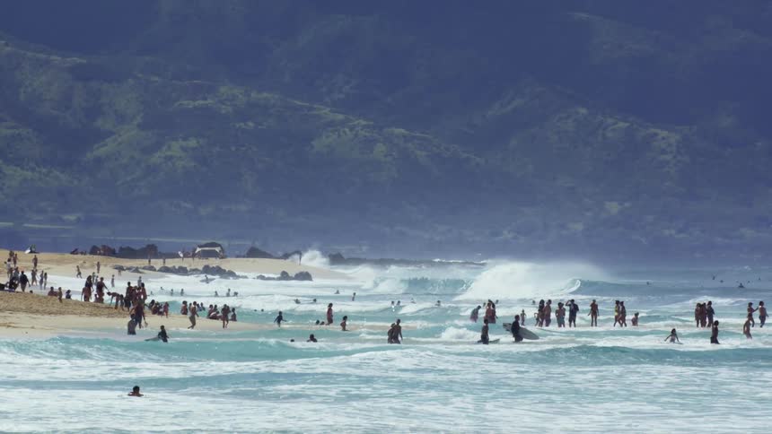 Dozens of people scattered across choppy turquoise waves near a sandy beach with powerful surf rolling in under a moody sky and lush green mountains rising in the background