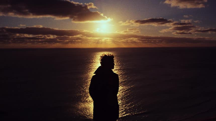A silhouetted person with curly hair stands on a cliff edge at dusk looking out over a calm ocean as the sun sets behind clouds casting a bright golden reflection across the surface of the water