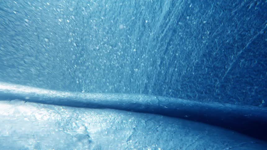 A camera moves through the interior of a breaking ocean wave capturing the turbulent blue water and white foam as the crest curls over to create a barrel filled with sunlight and thousands of tiny rising air bubbles