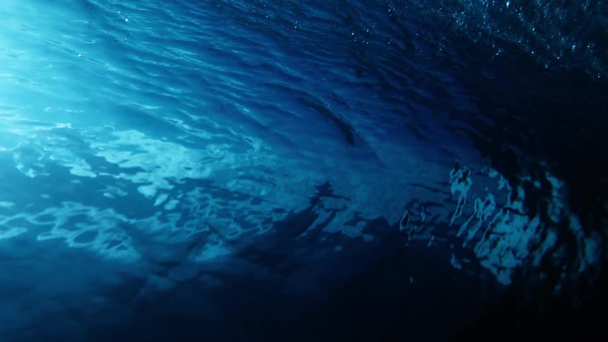 From an underwater perspective a breaking wave churns the surface while a surfer paddles through the dark blue water silhouetted against bright rays of sunlight piercing through the foam and bubbles