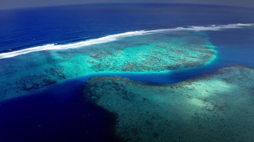 An aerial drone shot captures a vast coral reef system with vibrant turquoise shallows transitioning into deep blue ocean water under a clear sky showing waves breaking against the outer edge of the reefs natural limestone structure