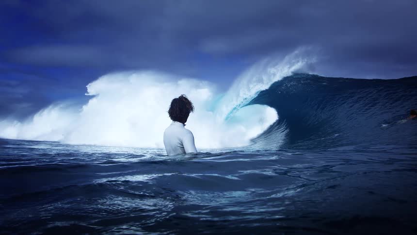 A surfer in a wetsuit floats in dark rolling water facing a towering curling blue wave beneath a dramatic stormy sky while another person stands in the water