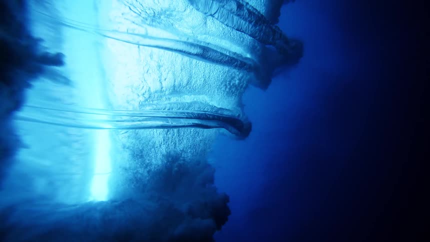 An underwater camera captures the churning white foam and swirling air bubbles of a large wave breaking overhead in the deep blue ocean as sunlight filters through the surface