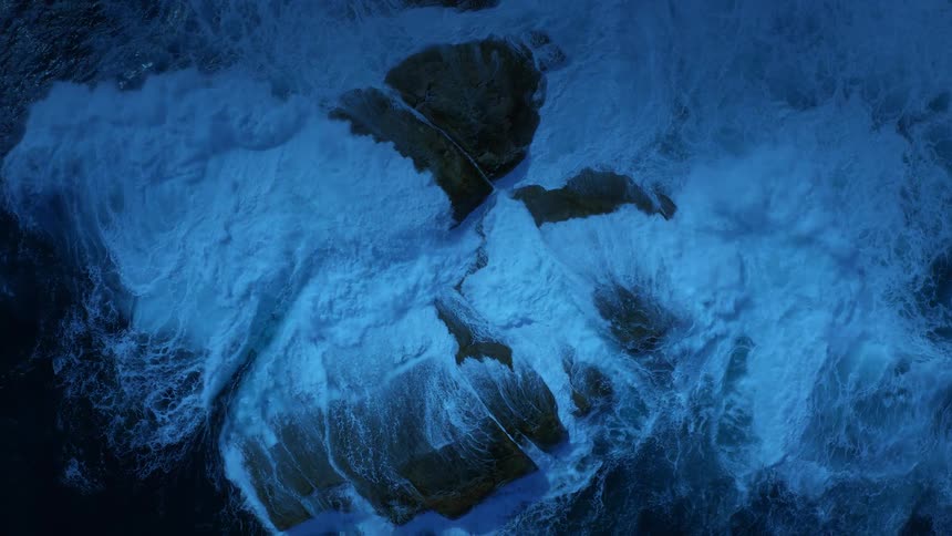 In this highangle aerial shot heavy blue waves surge and crash violently against a cluster of large dark rocks in the middle of the ocean creating a dense field of white foam and turbulent sea spray that completely swirls around the stones