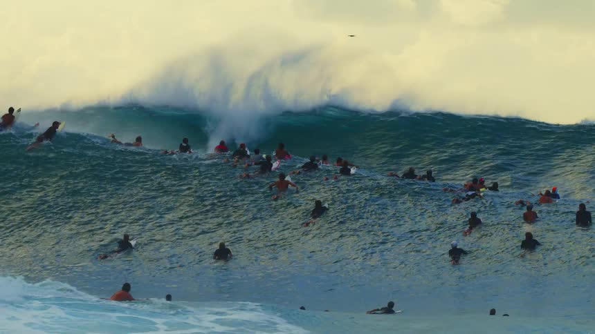 A wide shot shows a large crowd of surfers in the water as a massive powerful wave breaks behind them sending a high spray of white mist into the air several small drones are visible flying in the sky above the surfers