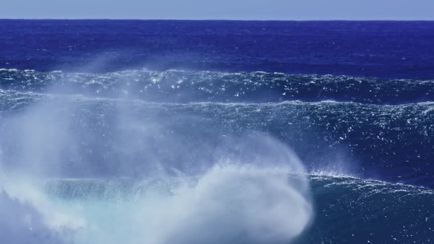 A powerful ocean wave crests and sprays mist under clear blue daylight