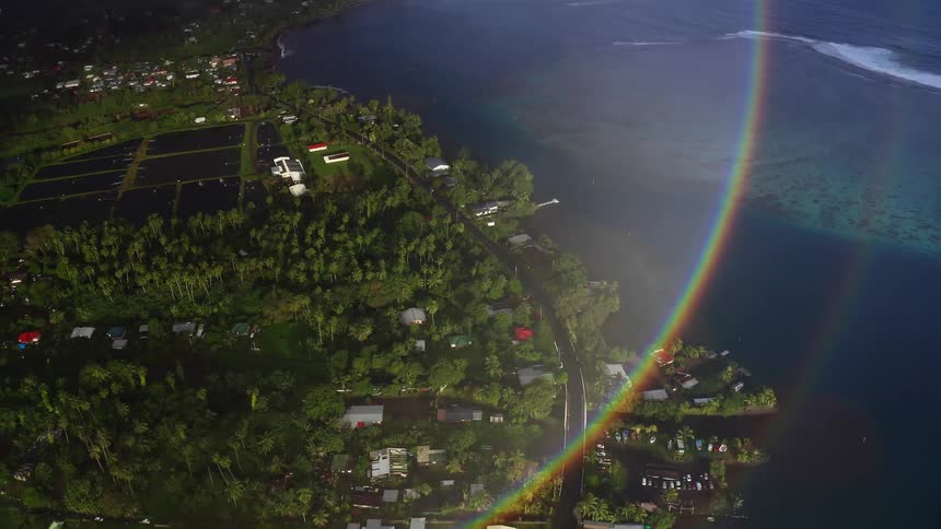 N aerial view reveals a lush tropical coastline dotted with small homes and winding roads while a vivid rainbow arcs across the frame over the shoreline and calm reflective water