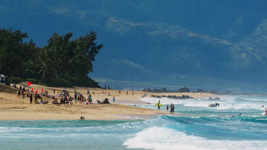 A scenic beach is filled with people relaxing on the sand and surfers in the water under a clear blue sky in the background lush green mountains rise against the horizon