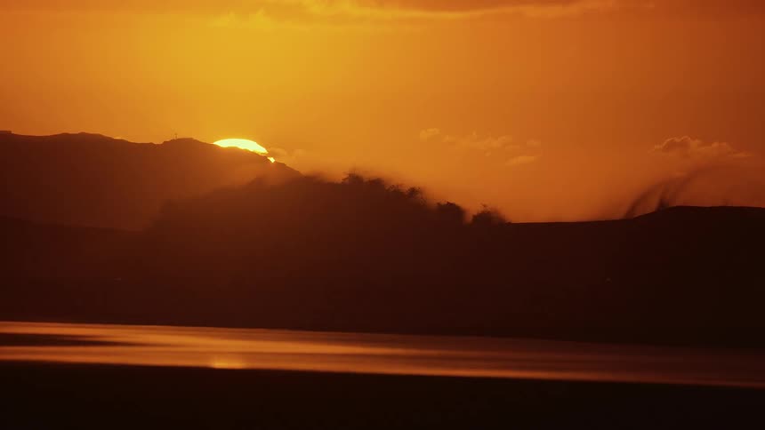 Silhouette of the mountain range in the midground it creates a dark uneven boundary against the bright orange sky with the sun partially tucked behind one of the peaks