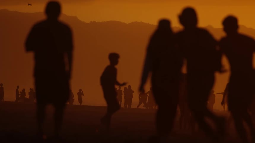 Silhouetted figures of adults and children move across a beach at dusk their dark outlines set against a warm orange sky with distant mountains