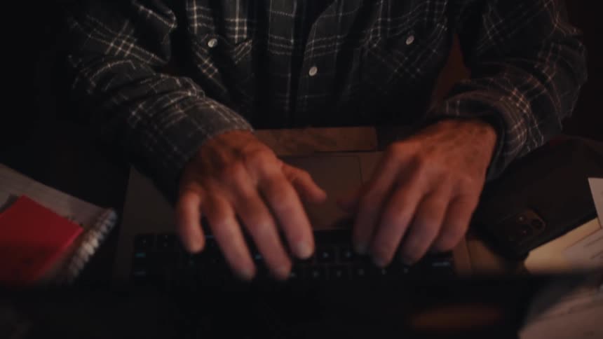 An elderly man typing on a laptop while seated indoor at night