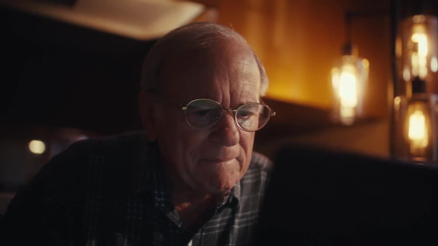 An elderly man sits at a table and concentrates on his work