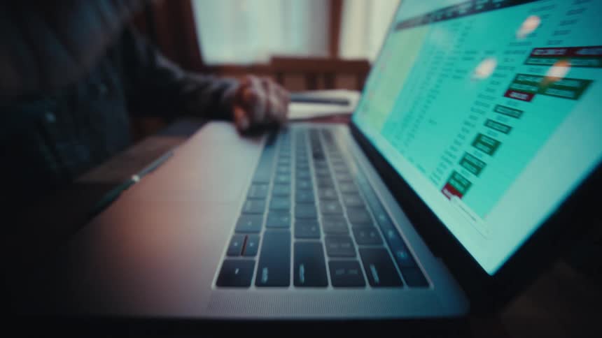 A man sits at a wooden table in a dimly lit room focusing on a laptop screen that displays a colorful spreadsheet of financial data and market figures