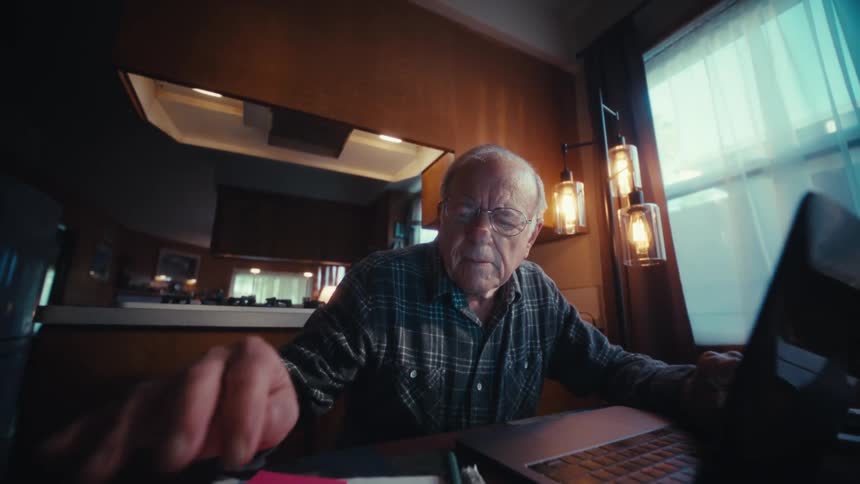Elderly man seated at a table using a calculator and a laptop to organize financial records in a home setting