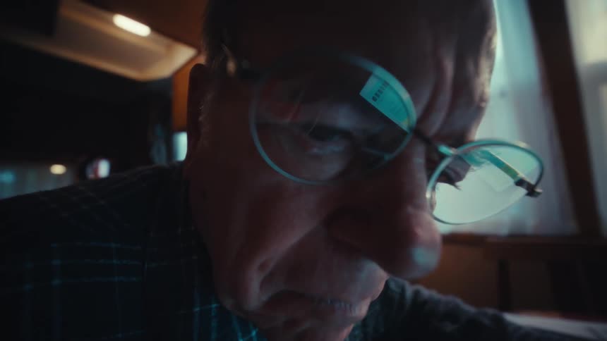 An elderly man sitting at a table uses a calculator and laptop