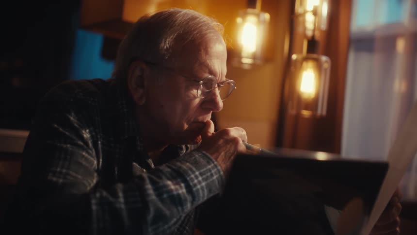 Elderly man sitting indoor at night holds a pen and a white paper with an opened laptop