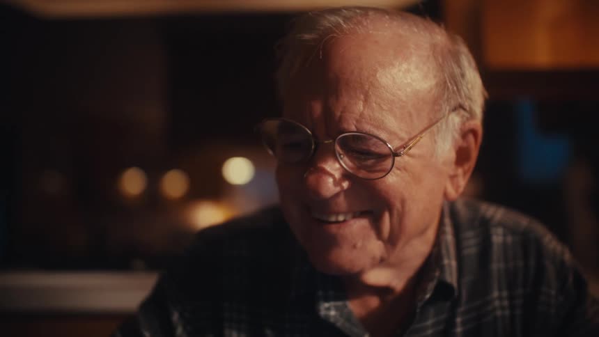 An elderly man happily works on a calculator at his desk in a dim indoor workspace