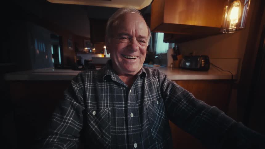 Elderly man wearing a plaid shirt with a visible smile in a home environment with a kitchen in the background