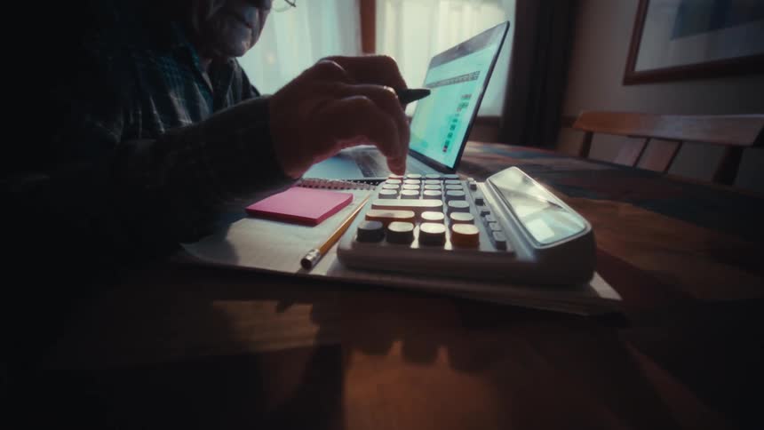 Man sitting at a desk looking at a laptop computer and pressing buttons on a calculator