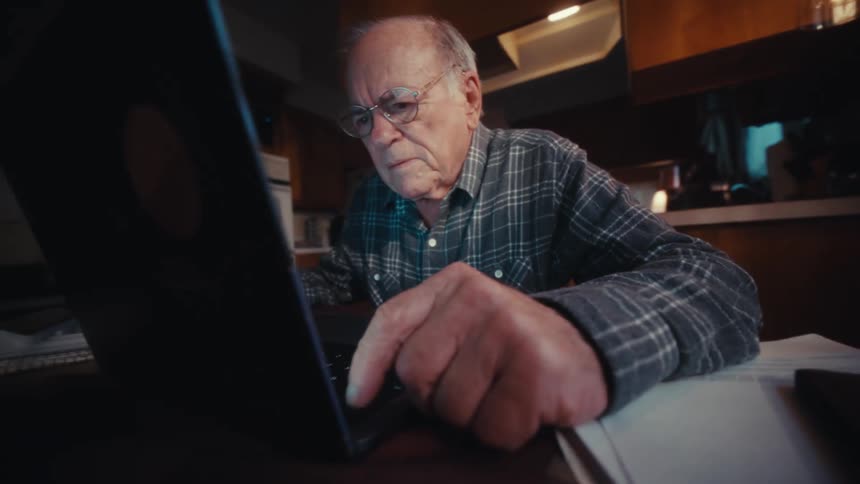 Elderly man concentrates while working on a laptop at a desk in a dim home workspace