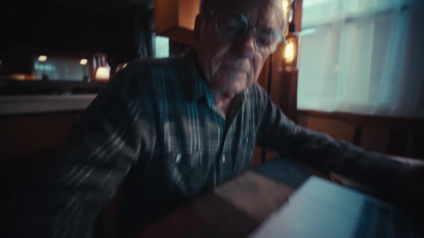 An elderly man works on a calculator while using a laptop in a dim home workspace