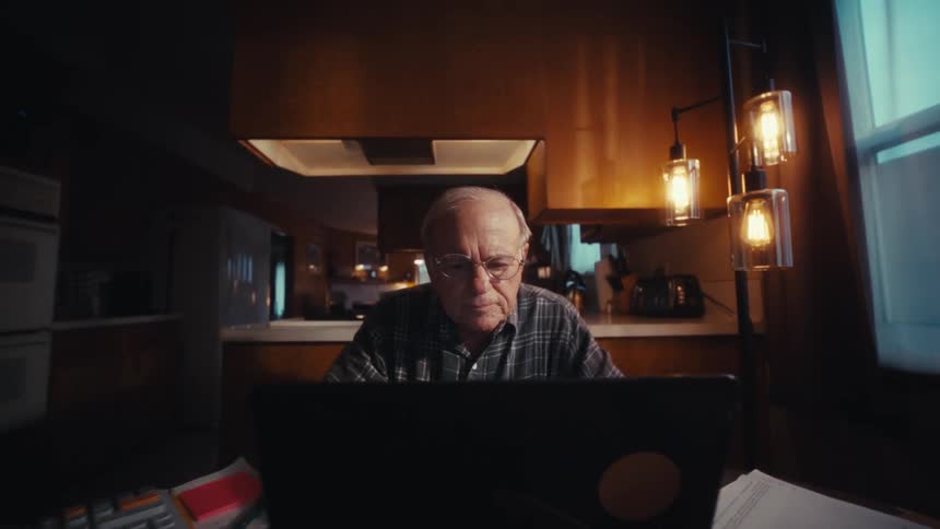An elderly man with glasses sits at a dimly lit kitchen table carefully reviewing papers and typing on his laptop beside a calculator as warm hanging lights illuminate his focused expression in the quiet evening