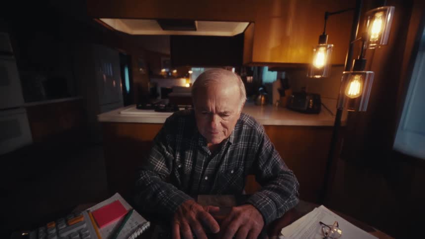 Elderly man sitting at a kitchen table with a calculator in the evening types on a laptop