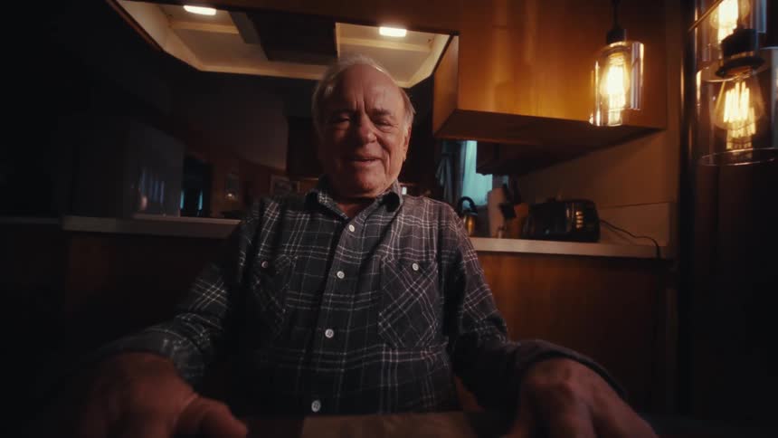 An elderly man relaxes and smiles after closing a laptop while sitting indoor at night