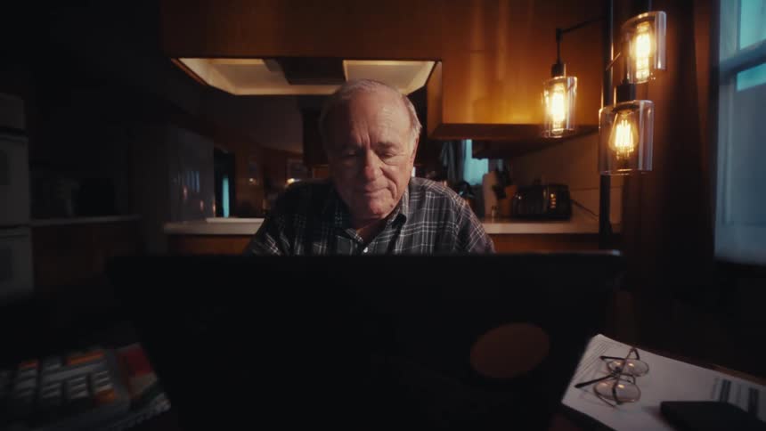 Elderly man seated at a table using a laptop with a calculator documents and a phone arranged nearby in a home environment