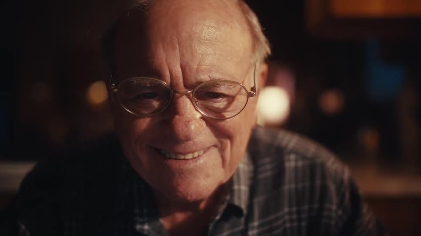 Elderly man wearing glasses with a slight smile in a warmlit indoor environment