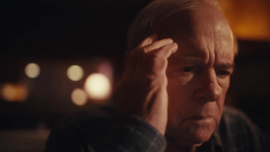 An elderly man sits in a dimly lit room at a desk with a laptop holding a paper as he appears nervous in thought