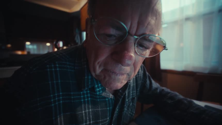 The video shows an elderly man in a plaid shirt focused on calculating finances at a desk he is seen alternating between using a calculator typing on his laptop and carefully reviewing documents