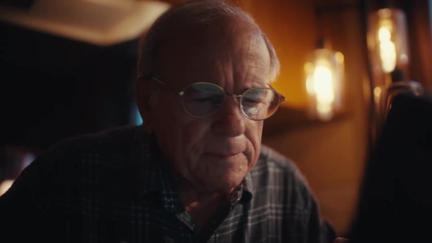 Elderly man sitting down at a table in front of a laptop computer on a living room and smiling indoors during day time