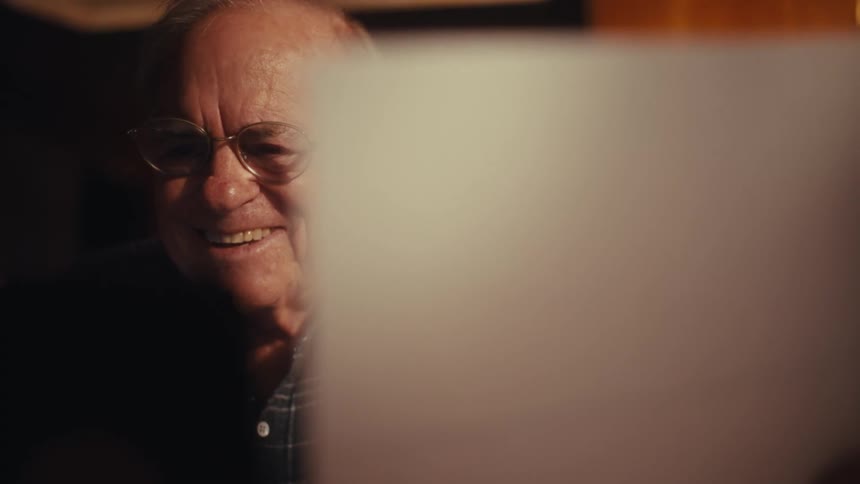 An elderly man wearing glasses and a plaid shirt reads a piece of paper and he begins to smile and laugh warmly before setting the paper down and resting his chin on his hand