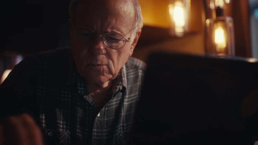 An elderly man wearing glasses and a plaid shirt works on a laptop computer at a desk in a dimly lit room with warm ambient lighting