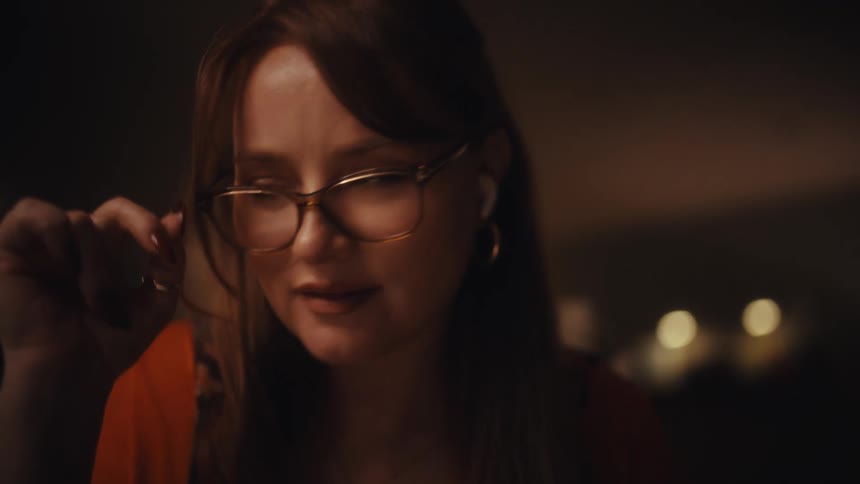 A woman wearing glasses and wireless earbuds looks downward with a thoughtful expression