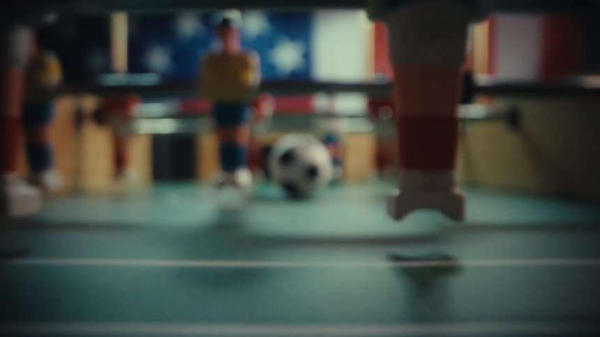 A closeup shot of a black and white foosball ball sits on a green table before the camera pulls back through the goal hole to reveal the plastic players and the dark room setting