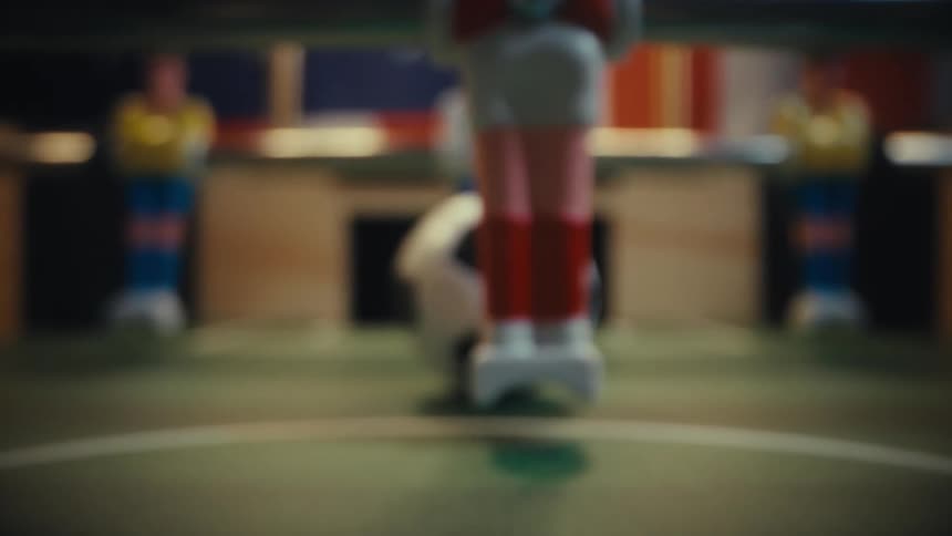 A closeup shot moves across a foosball table following a small soccer ball as it rolls toward the center of the pitch while the plastic players on rotating metal rods blur in the foreground and background during a game