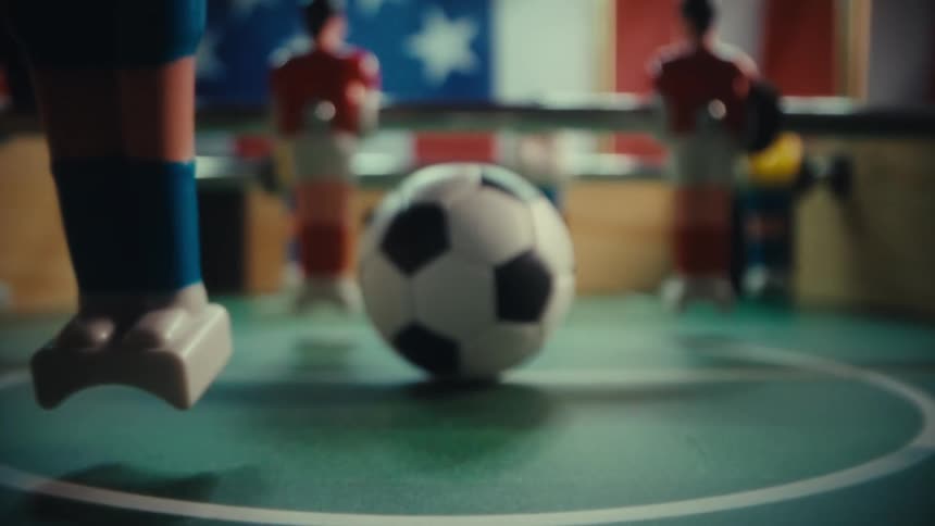 The camera moves forward along the surface of a foosball table passing between plastic toy players to focus on a miniature black and white soccer ball sitting stationary in the center of the green pitch under bright indoor lighting