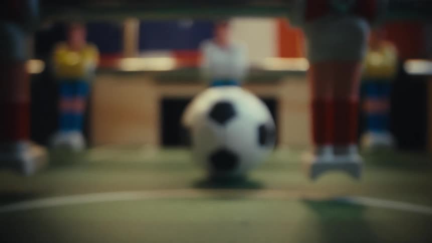 A foosball figures legs beside a blackandwhite ball at center field while blurred opposing players and a dimly lit game room in the background