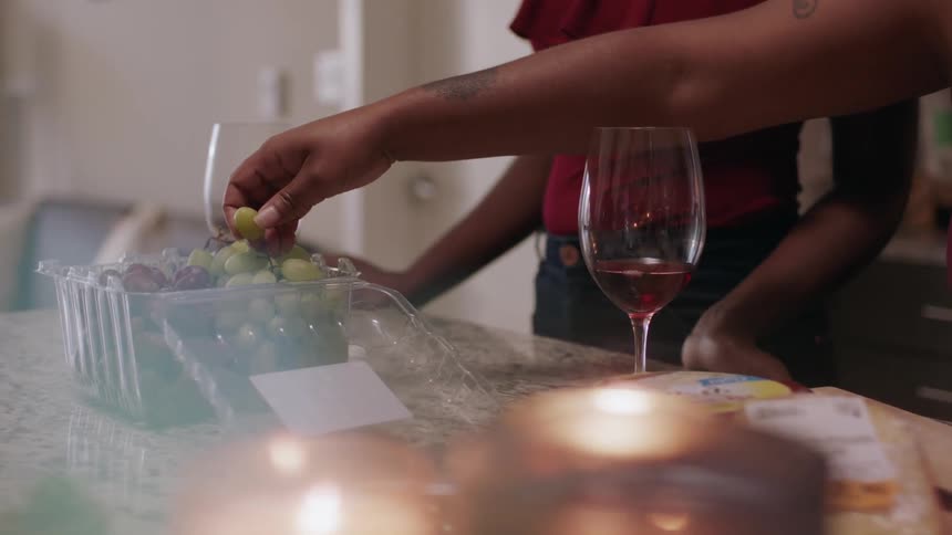 Two people at a kitchen counter appear to be having wine and grapes