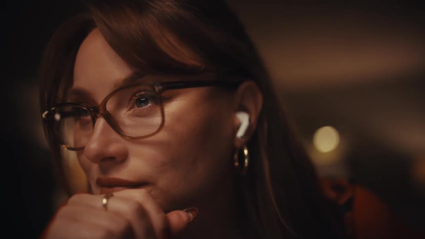 A woman wearing glasses and wireless earbuds adjusts her frames and rests her chin on her hand with a thoughtful expression